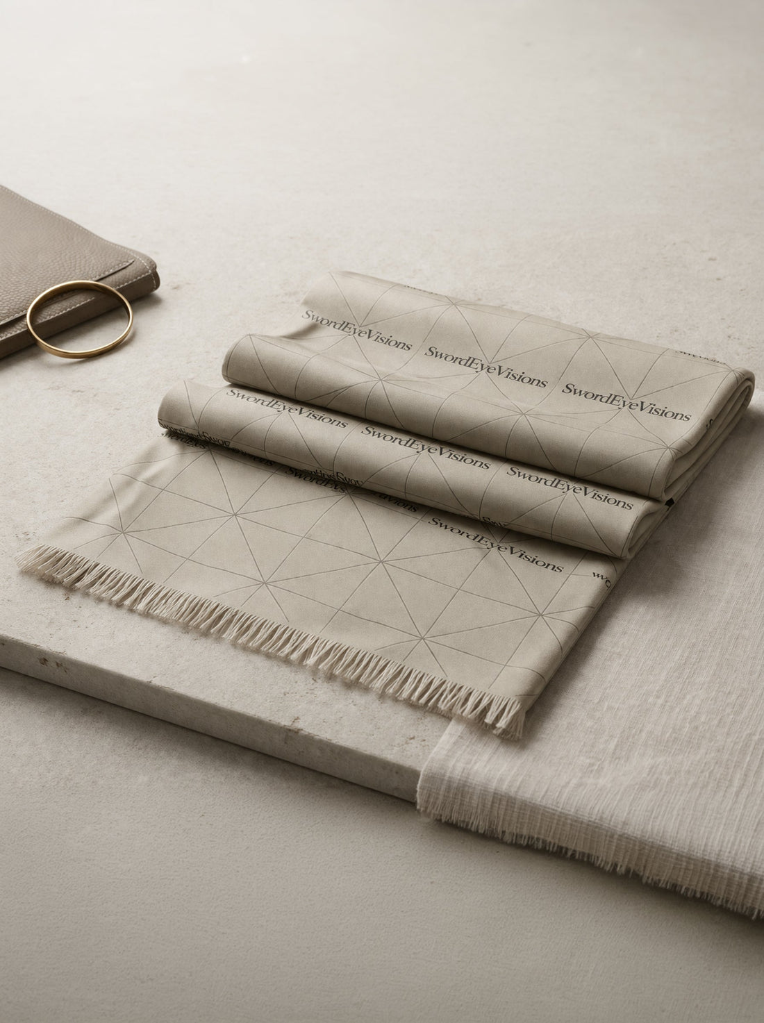Folded neutral pebble stone SwordEyeVisions scarf with tessellated linear pattern on a stone surface.
